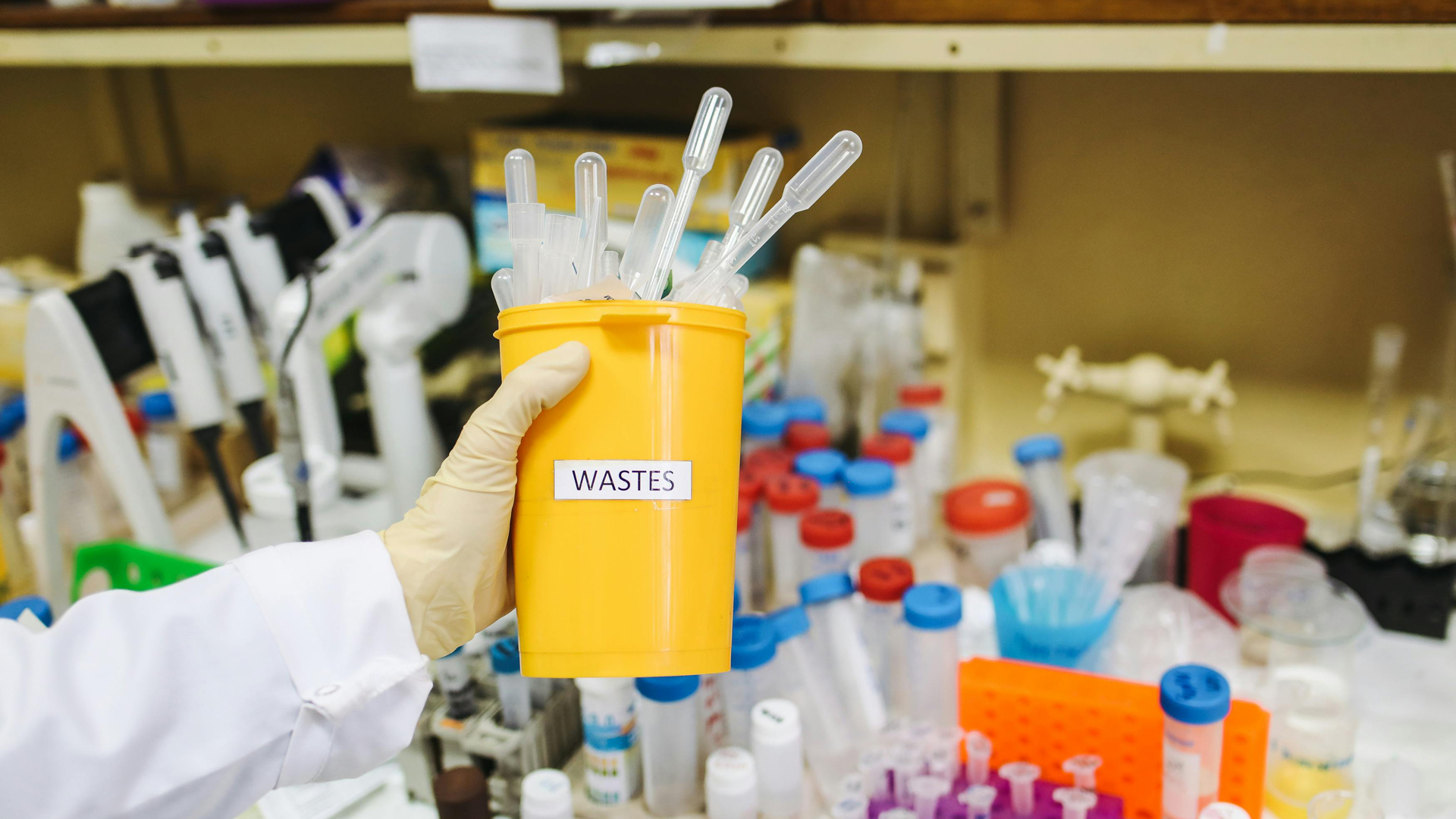 An arm and hand wearing a lab coat and rubber glove holding a yellow container labelled waste full of plastic liquid droppers infron of medical research tubes and containers